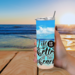 Woman holding tumbler with Life Is Better quote; her hand on table, beach scene in background, relaxing ocean vibes.